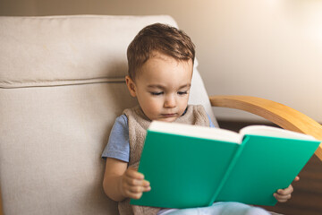 A little boy is sitting on a chair and reading a book. Toddler. Happy childhood. Reading books for...