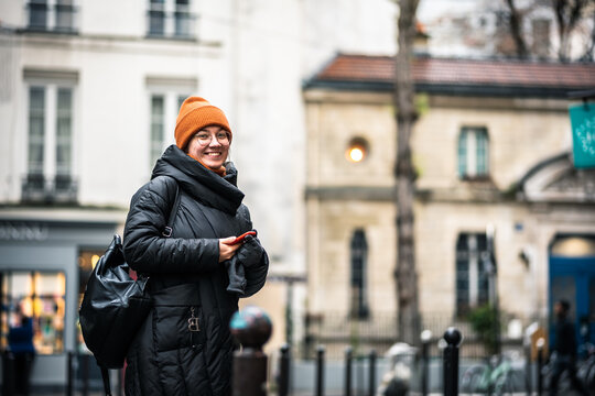 Happy smiling woman walking in Paris city in winter. Old city. Tourist. The girl is standing with the phone. The girl is wearing a black coat and an orange hat. Walking down the street.