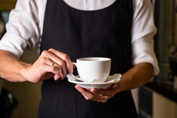 barista in a white shirt and black apron holds a cup of coffee