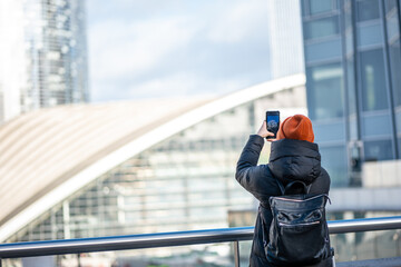 Young Woman Photographing Modern Glass Skyscrapers in Black Winter Coat and Orange Hat 