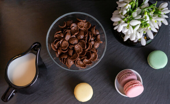 Breakfast with chocolate cereals, milk, macaroons,  pink heart shaped chocolate pralines and snowdrops in a black vase, on a black background