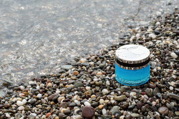 Fototapeta na wymiar Skin care cream in a blue glass jar with a shiny silver cap on the seashore in pebbles, with water