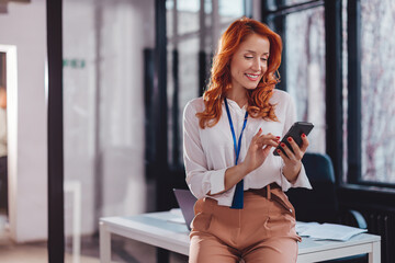 Beautiful young businesswoman using mobile phone in office while leaning on desk.