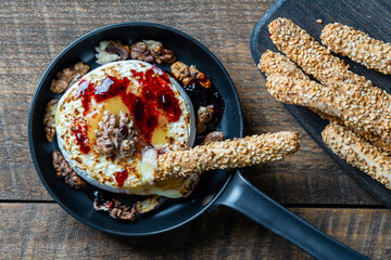 View of baked on frying pan camembert cheese, walnuts, honey, jam and bread sticks with sesame seeds on wooden table background, closeup, top view