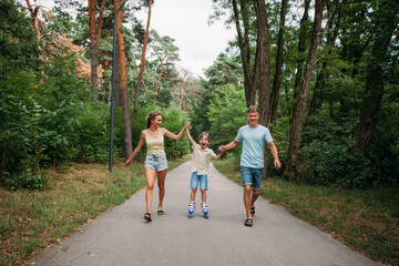 A happy young family, dad, mom and son are skating hand in hand with their parents, on roller skates, along a path in the park. The whole family is dressed in shorts and T-shirts.