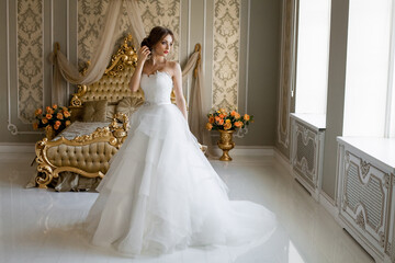 A beautiful smiling bride with chic makeup and hairstyle in a white wedding dress is standing in a bedroom with a stylish light vintage interior, in the morning before the wedding.