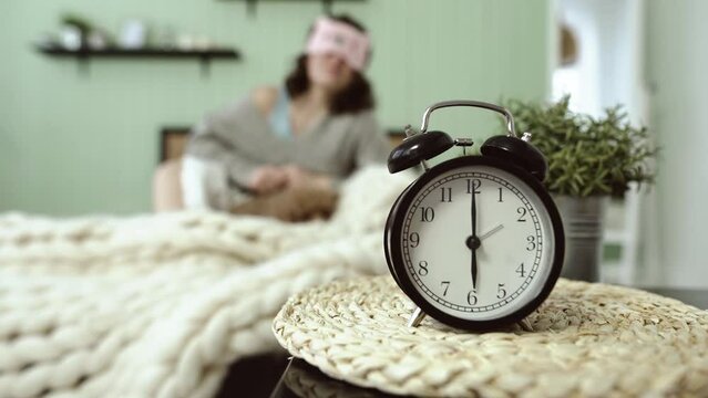 Six a.m. on alarm clock, in background a young woman wakes up on bed