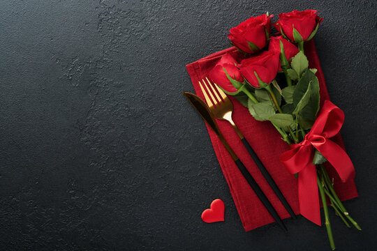 Valentines day romantic table setting. Empty closeup of red roses, wine, candles, dinner black plate, knife, fork and decorative silk hearts on black background. Holiday concept. Copy space. Top view