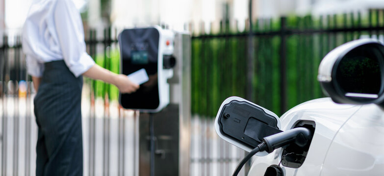 Focus Electric Vehicle Recharging At Public Charging Station With Blurred Progressive Woman Using Credit Card Payment For Electric Refuel For Her EV Car For Alternative Eco-friendly Car Concept.