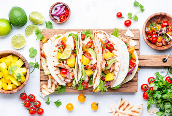 Tacos with grilled chicken, tomato salsa sauce, mango, cilantro and onion in tortilla on cutting board. White table background, top view