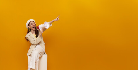 Young Asian woman sitting on white box holding megaphone and pointing finger isolated on yellow background. Speech and announce concept.