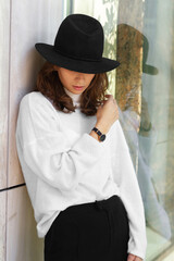 Young brunette sexy girl woman in a romantic look in a white winter cosy sweater and black pants and hat in an urban city center near the glass wall with plants.