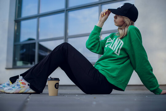 Beautiful Girl Model With Coffee In A Black Jacket In A Black Baseball Cap On A Chair Sitting Outside On The Stairs Posing For The Camera With A Phone In Green Clothes In Sneakers Fashion