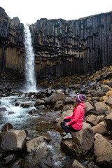 Girl in pink jacket looking at powerful stunning famous svartifoss waterfall in picturesque scenery...
