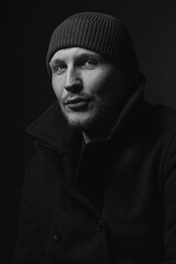Naklejka na ściany i meble Lifestyle, fashion, occupation concept. Man with winter hat and black coat studio portrait. Model with beard looking at camera with serious look. Dark studio background. Black and white image