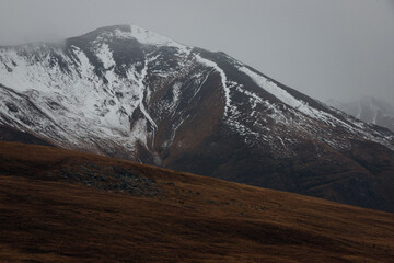 Photo of the slope of hill and the peak of mountain covered snow in Autumn foggy and cloudy day. 