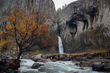 Photo of the waterfall named Karakaya-Su that falls in the canyon from the cleft of rocks from 30 meters. It was Autumn cloudy day, on mountain riverbank there is a tree with yellow leafs.