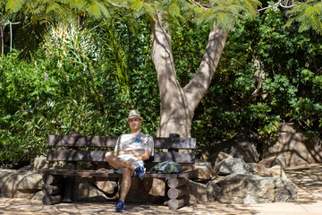 Turysta siedzi na ławce w cieniu drzewa - obrazy, fototapety, plakaty