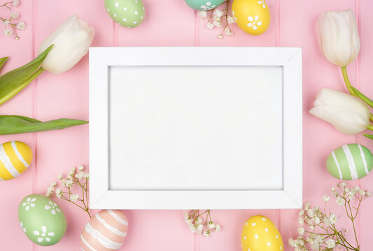 White wood frame with pastel Easter eggs and white spring tulip flowers. Above view against a pink wood background. Copy space.
