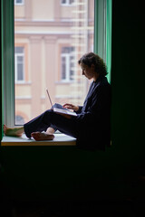 Profile shot of female in business suit looking at laptop during working process sitting on the window in co-working open office space on the dark background. High quality photo