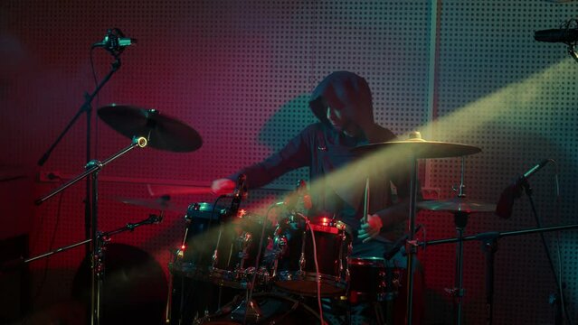 Drummer performs at percussion instruments at rock concert. Tympanist man plays drums, musical cymbals at show. Musician in hoody at rehearsal, participates in recording of album in sound studio