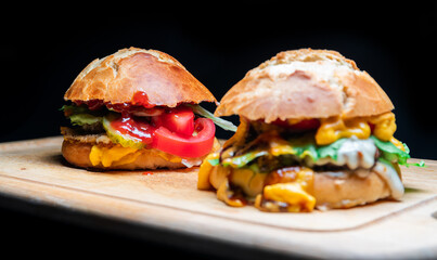 Burger mit Brötchen, Soße, Zwiebeln, Salat, Tomate, Käse auf einem Holzbrett vor einem schwarzen Hintergrund