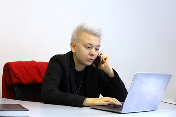 Girl with short blonde hair sitting at laptop and talking on mobile phone. Tomboy lifestyle, concept of office work, business assistant