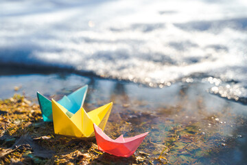 Multicolored paper boats. Colorful pink yellow blue orange ships in big spring snow puddle on winter street. Warm wet rainy weather, old grass. Hello spring, autumn. Children play, have fun outdoors