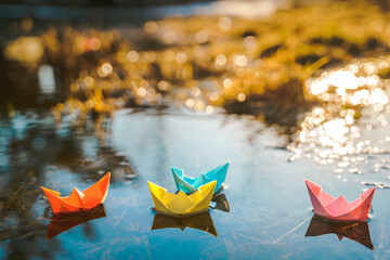 Multicolored paper boats. Colorful pink yellow blue orange ships in big spring snow puddle on winter street. Warm wet rainy weather, old grass. Hello spring, autumn. Children play, have fun outdoors