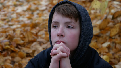 A hooded boy crossed his arms in front of his face against the backdrop of autumn foliage. Portrait of a sad and pensive teenage boy in nature.