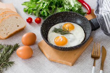 Fried eggs in a frying pan with rosemary, bread, eggs and green salad on a linen tablecloth. Breakfast, lunch and dinner. The view is soft, selective focus. Rustic style.