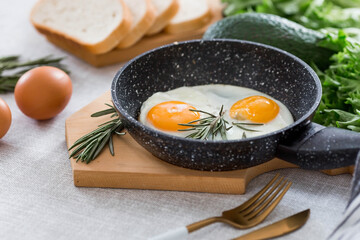 Fried eggs in a frying pan with rosemary, bread, eggs and green salad on a linen tablecloth. Breakfast, lunch and dinner. The view is soft, selective focus. Rustic style.