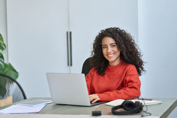 Young happy pretty latin business woman company worker sitting at desk working on laptop. Smiling female professional executive using computer in corporate office looking at camera. Portrait. - obrazy, fototapety, plakaty