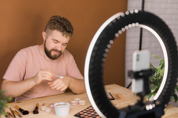 Male beauty blogger with makeup product recording video at home. Using ring lamp and smartphone....