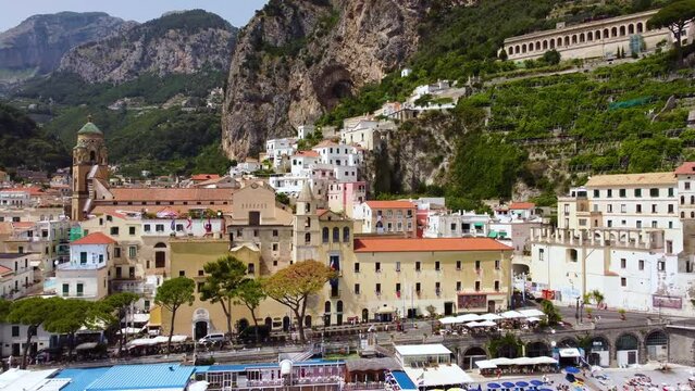 Aerial drone footage of Marina Grande beach, town hall, Sant'Andrea duomo bell tower in Amalfi coast, Campania, Italy. Parasol, sun loungers, people swimming and colourful villas on cliff from above.