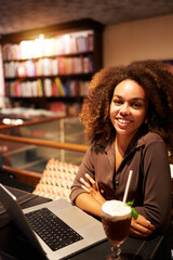 portrait smiling young afro black brazilian woman sitting in a library cafeteria with a notebook...