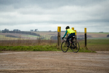 a sports cyclist wearing a yellow hig-vis top, riding a racing bike past tank crossing marker posts