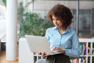Young successful confident African American woman entrepreneur, CEO or office worker working using laptop, stands in modern office or coworking
