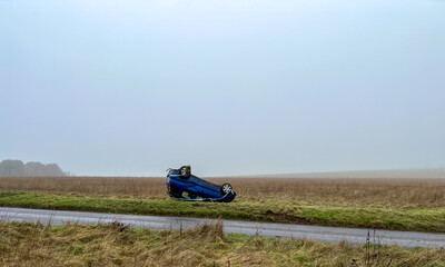 an overturned blue Suzuki hatchback car resting on its roof having crashed off a road on to grass
