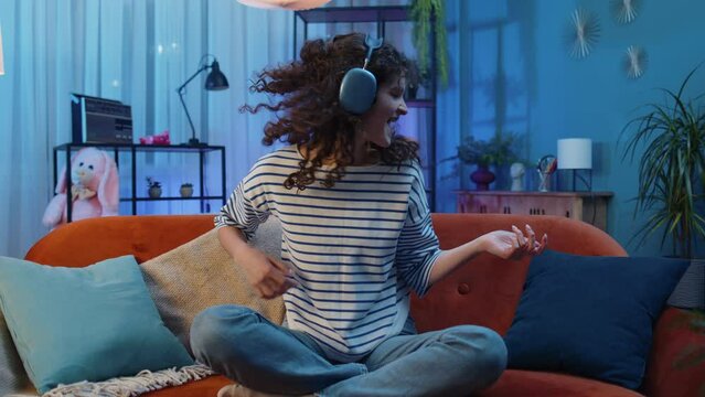 Happy rocker woman with curly hairstyle in wireless headphones relaxing at home dancing on couch listening energetic disco dancing music, playing on imaginary guitar. People weekend leisure activities