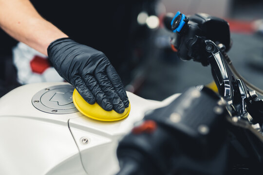 Hand in a black protective glove using delicate sponge to clear and polish the surface of white motorcycle. Closeup shot. Copy space. High quality photo