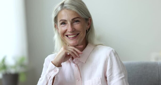 Optimistic middle-aged 50s blond woman smile looking at camera, sit on sofa, enjoy relaxation, feels carefree on weekend alone at home. Beautiful mature homeowner female close up, head shot portrait