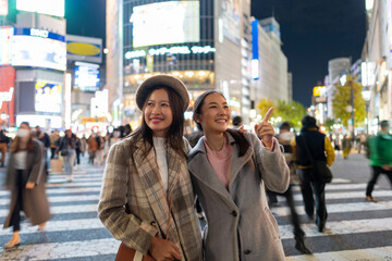 Asian woman shopping and crossing street crosswalk with crowd of people at Shibuya, Tokyo, Japan at night. Happy girl friends enjoy and fun outdoor lifestyle travel in the city in autumn vacation.