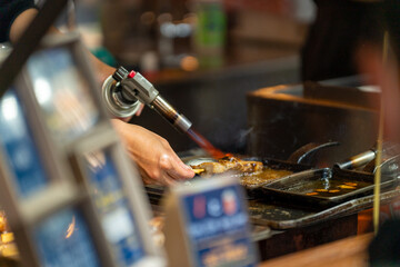 Professional male chef hand burning street food grilled tuna on stick serving to customer at fish market in Tokyo city, Japan. Japanese street food and street market travel vacation concept.
