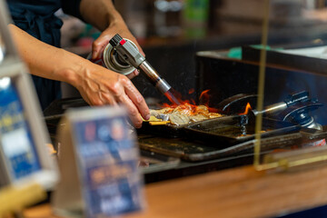 Professional male chef hand burning street food grilled tuna on stick serving to customer at fish market in Tokyo city, Japan. Japanese street food and street market travel vacation concept.
