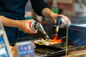 Professional male chef hand burning street food grilled tuna on stick serving to customer at fish market in Tokyo city, Japan. Japanese street food and street market travel vacation concept.
