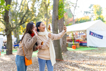 Attractive Asian woman friends enjoy and fun using mobile phone taking selfie together while eating street food together at outdoor festival in Tokyo city, Japan in autumn holiday vacation. 