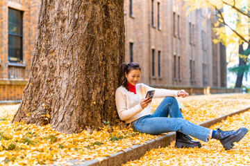 Happy Asian woman using mobile phone while travel outdoor at park in Tokyo city, Japan on holiday vacation. Attractive girl looking beautiful nature of yellow ginkgo tree leaves falling down in autumn