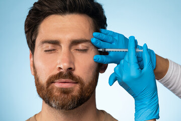 Handsome middle aged bearded man getting under eye injections in beauty clinic, blue studio...