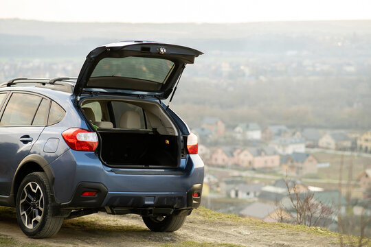 Blue Off Road Car With Open Trunk Lid At Dawn On Grassy Hill On Distant Foggy City Landscape Background